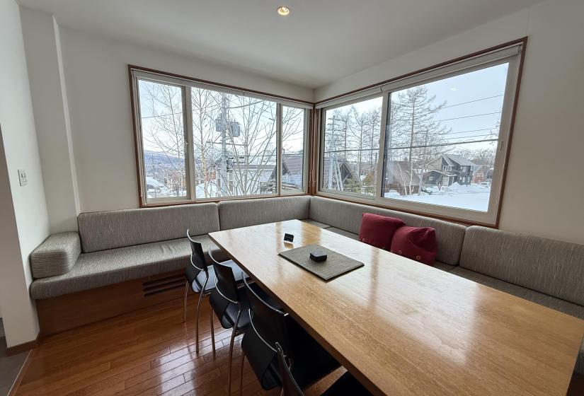 Bright dining area at Yutaka Townhouses with wooden dining table, bench seating with red cushions, corner banquette, and panoramic windows offering stunning views of snowy Niseko forest and mountains, perfect for family meals during ski holidays.