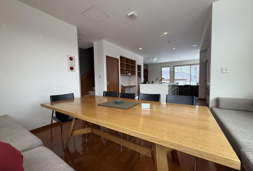 Spacious open-plan dining and living area at Yutaka Townhouses showcasing wooden dining table, kitchen with modern appliances, and comfortable living space beyond, offering convenient social layout for groups enjoying Niseko ski holidays.