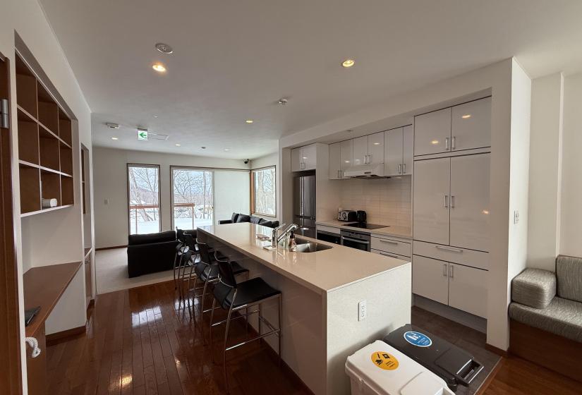 Spacious modern kitchen at Yutaka Townhouses featuring white cabinetry, large central island with bar seating, sleek countertops, full-sized appliances, wooden flooring, and bright windows, offering premium self-catering facilities for Niseko ski holiday guests.