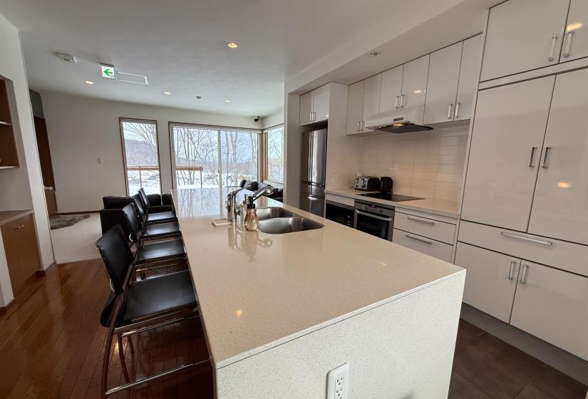 Kitchen and dining space at Yutaka Townhouses showcasing expansive island with bar seating, sleek white cabinetry, modern appliances, and open-plan layout flowing to living area, perfect for social gatherings during Niseko ski holidays.