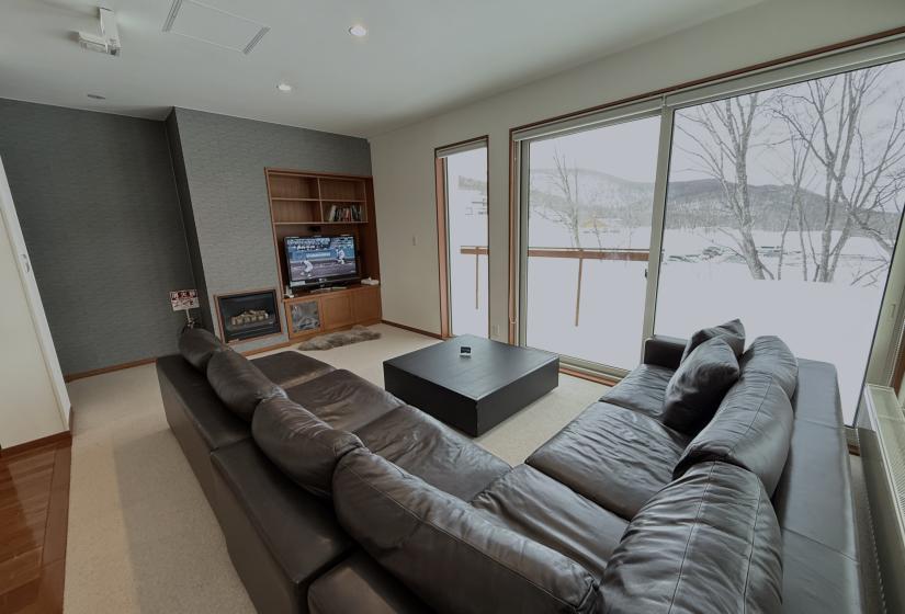 Comfortable living room at Yutaka Townhouses with plush gray sectional sofa, flat-screen TV, wooden media console, floor-to-ceiling windows overlooking snowy Niseko landscape, and contemporary design perfect for relaxing after a day on the slopes.