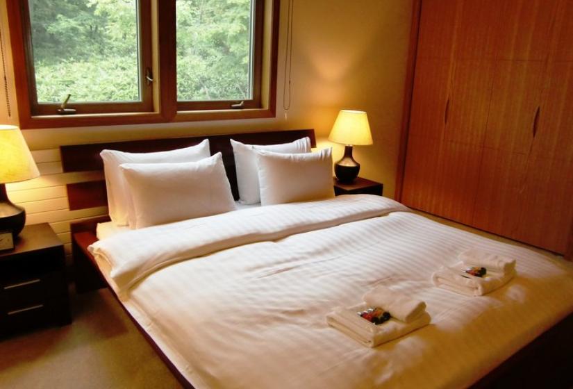 Bedroom with double bed featuring white linens, wooden wall paneling, large windows with forest views, bedside tables with lamps, and folded towels on the bed at Chalet Creekside.