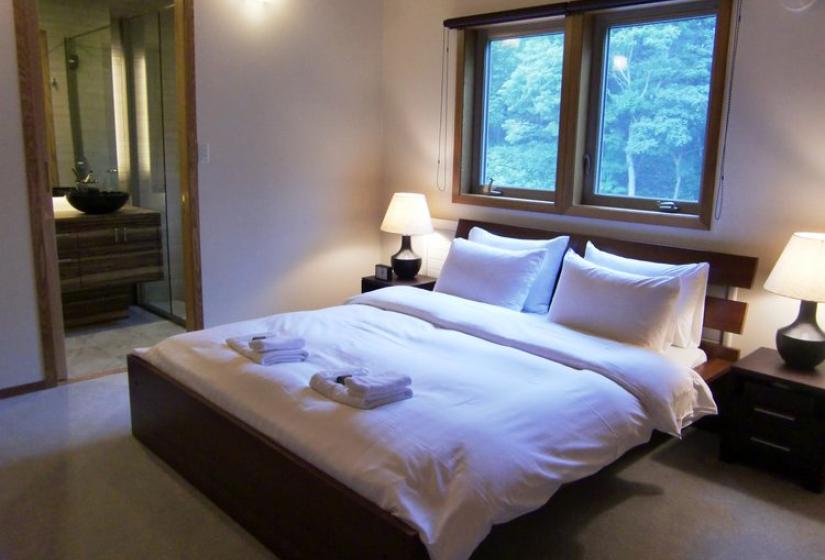 Queen bedroom with white bedding, wooden window frames with forest views, bedside tables with lamps, and an entrance to an en-suite bathroom at Chalet Creekside.