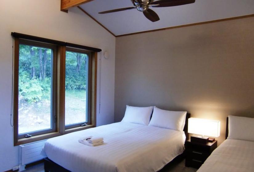 Bedroom with double bed featuring white linens, wooden window frames with forest views, ceiling fan, and bedside lamp at Chalet Creekside.