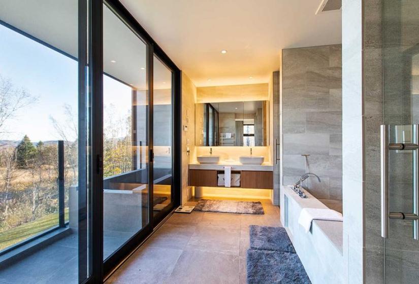 Luxurious bathroom in premium Niseko ski chalet featuring a modern soaking tub, expansive floor-to-ceiling windows overlooking birch forest, elegant stone tiles, glass shower enclosure, and contemporary vanity with backlit mirror - perfect for relaxation after skiing Japan's legendary powder.