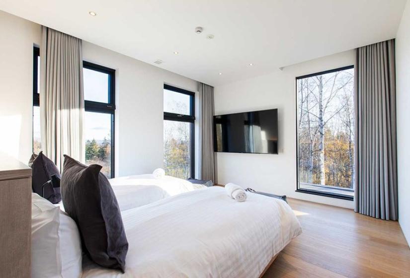 Luxurious bedroom in upscale Niseko ski chalet featuring a plush king bed with premium white linens, sleek wall-mounted TV, contemporary furnishings, floor-to-ceiling windows with forest views, and elegant gray curtains - offering refined comfort after days on Japan's legendary powder slopes.