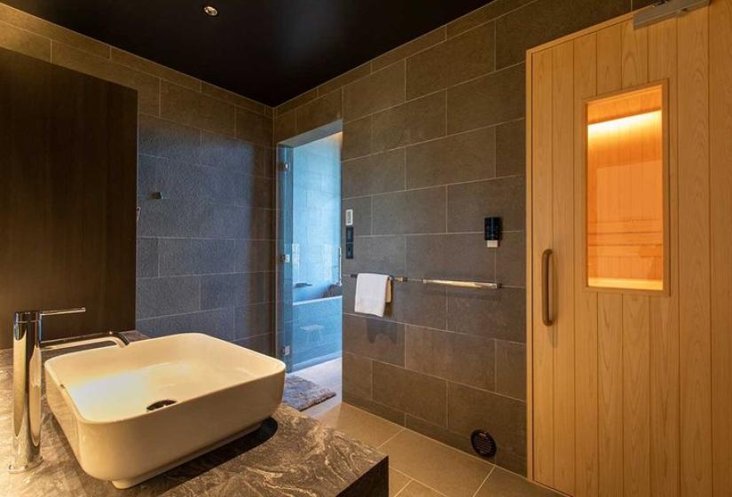 Luxury bathroom in Niseko ski chalet featuring a contemporary freestanding bathtub, elegant stone tile walls, large windows with mountain views, and a private sauna with warm wood interior - perfect for après-ski relaxation in Japan.