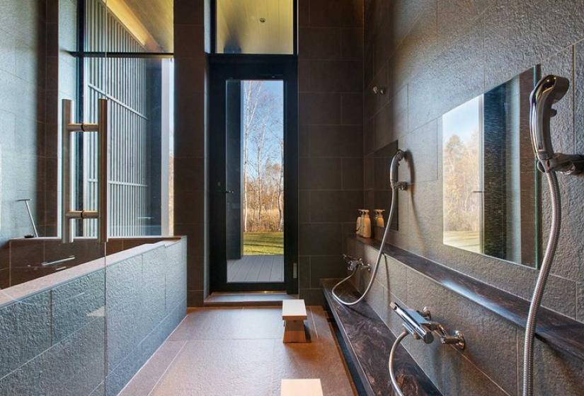 Spacious luxury shower room in Niseko ski chalet featuring dual rainfall showerheads, premium stone tiling, large windows with forest views, and minimalist Japanese design elements for ultimate après-ski relaxation.