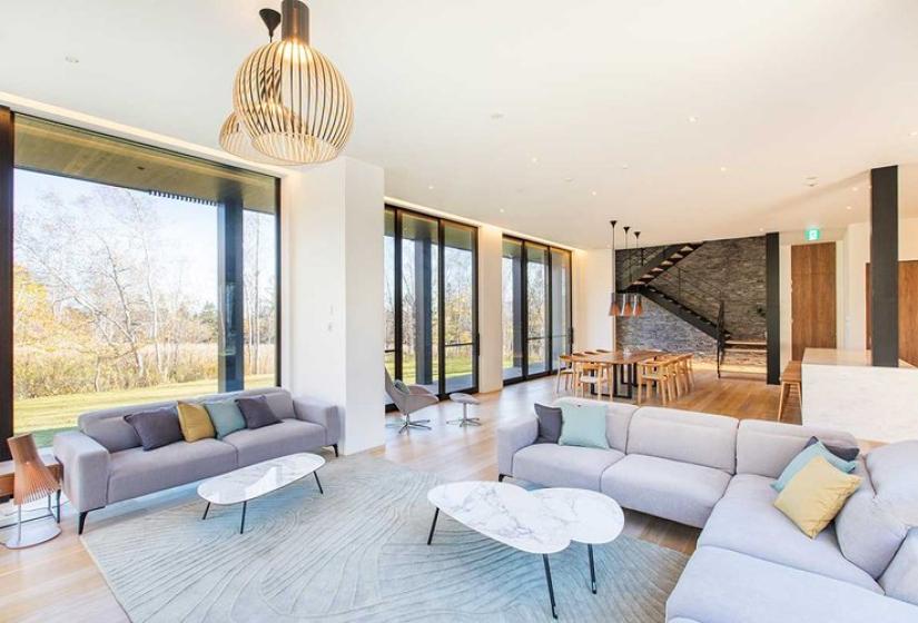 Contemporary open-concept living room in luxury Niseko ski chalet featuring comfortable gray sectional sofas, designer pendant lighting, stylish coffee tables, soft area rug, and expansive windows showcasing stunning forest views - providing an elegant gathering space after skiing Japan's world-famous powder.