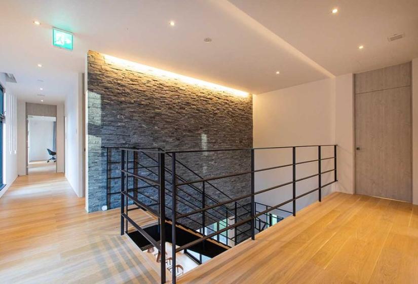 Contemporary Niseko ski chalet interior featuring an elegant upper hallway with wooden flooring, sleek black metal railings, and a dramatic stone feature wall with ambient lighting - representing premium Japanese alpine architectural design.