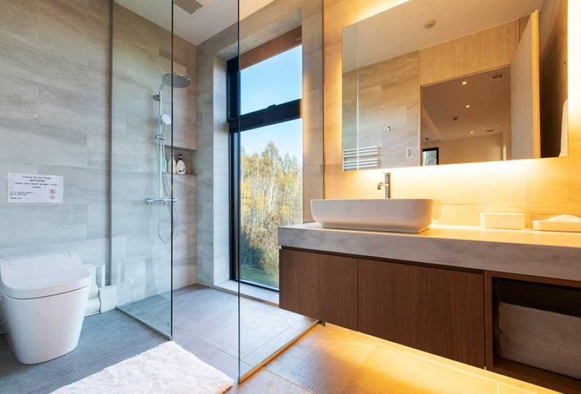 Sleek modern bathroom in luxury Niseko ski chalet featuring walk-in glass shower, elegant stone tile walls, floating dark wood vanity with vessel sink, backlit mirror with ambient lighting, and large window with forest views - offering stylish comfort after days on Japan's legendary powder slopes.