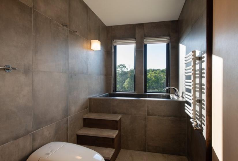 Elegant jacuzzi room in Chalet Kiraku featuring a built-in soaking tub with steps, contemporary concrete walls, large windows with forest views, and natural light, offering a serene post-skiing relaxation space for luxury accommodation guests in Niseko.