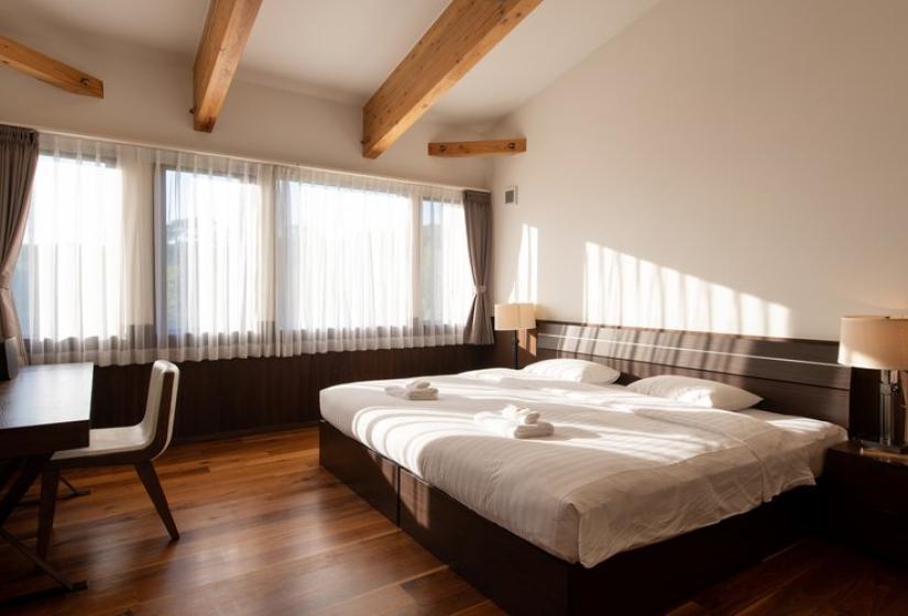 Bright and airy twin bedroom in Chalet Kiraku with exposed wooden beams, large windows with mountain views, comfortable beds with premium white linens, wooden flooring, and elegant Japanese-inspired furnishings, ideal for friends or family sharing luxury accommodation in Niseko.