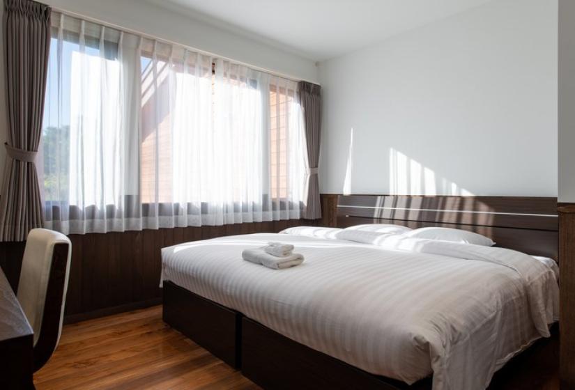 Spacious bedroom in Chalet Kiraku featuring a comfortable king-size bed with crisp white linens, wooden flooring, large windows with sheer curtains, and minimalist Japanese-inspired design, offering a peaceful retreat after skiing in Niseko.