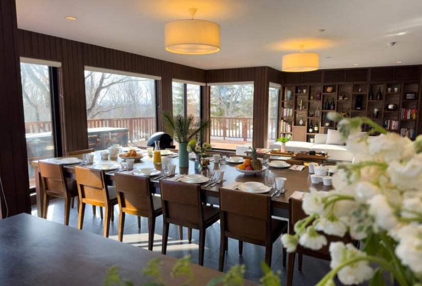 Spacious dining area in Chalet Kiraku featuring a long wooden table with comfortable seating for 12 guests, panoramic windows with mountain views, stylish pendant lighting, and fresh floral arrangements, perfect for luxury group stays in Niseko.