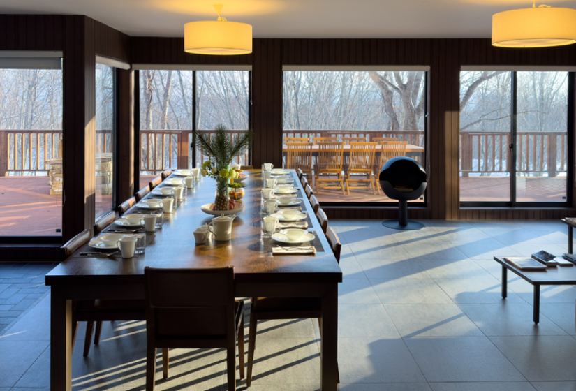 Elegant open-plan living and dining space in Chalet Kiraku featuring floor-to-ceiling windows with mountain views, a long wooden dining table, comfortable lounge seating, contemporary pendant lighting, and stylish decor, perfect for socializing during luxury ski holidays in Niseko.