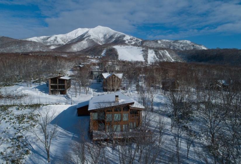 Aerial view of luxury chalets nestled in a snow-covered forest with Niseko ski resort slopes and mountains visible in the background, showcasing the exclusive location and pristine winter landscape surrounding this premium accommodation in Japan.