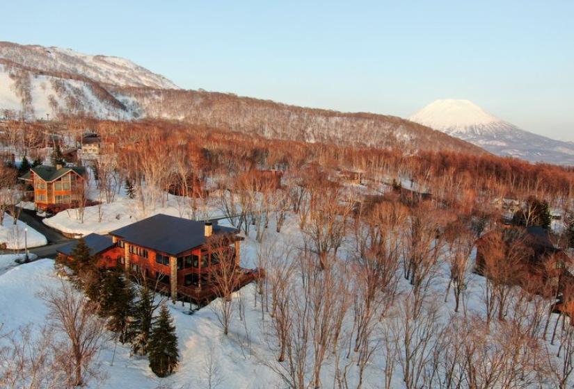 Stunning winter view of a luxury wooden chalet nestled among snow-covered trees in Niseko, with Mount Yotei visible in the background. Exclusive ski accommodation surrounded by pristine winter landscape and vibrant autumn foliage.