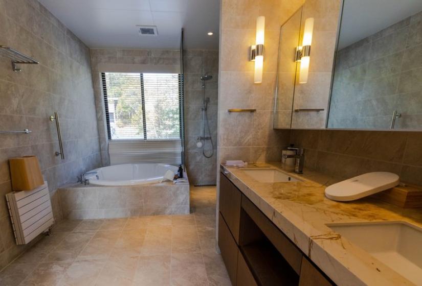 Luxurious master bathroom in Chalet Lotus Niseko featuring a freestanding white soaking tub, elegant stone walls, double vanity with vessel sinks, and warm lighting, offering spa-like comfort for guests.