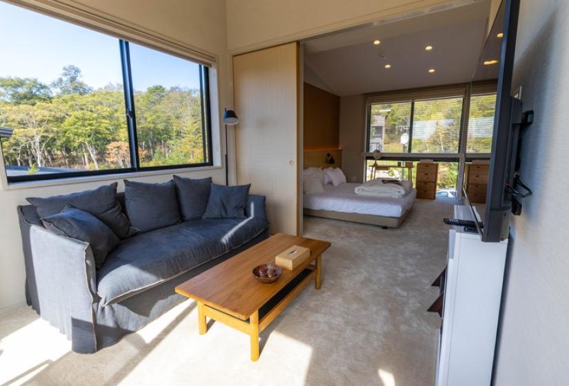 Spacious master bedroom suite at Chalet Lotus Niseko featuring a king bed, comfortable sitting area with dark sofa, wooden coffee table, floor-to-ceiling windows with stunning mountain views, and modern minimalist design.