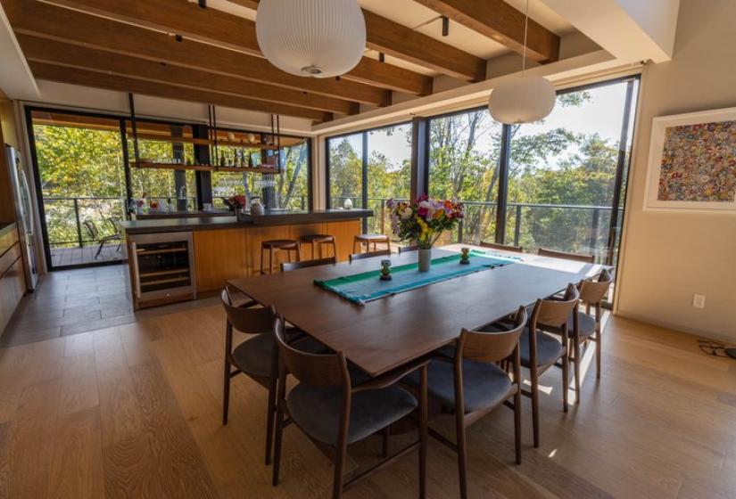 Bright open-concept dining and kitchen space at Chalet Lotus Niseko featuring a wooden dining table, contemporary chairs, exposed ceiling beams, paper lantern lighting, and expansive windows showcasing panoramic forest views for a seamless indoor-outdoor luxury experience.