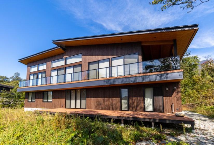 Modern two-story luxury chalet in Niseko featuring extensive glass windows, wooden exterior with dark trim, spacious decks, and contemporary architectural design set against natural surroundings, located minutes from Annupuri's renowned ski slopes.