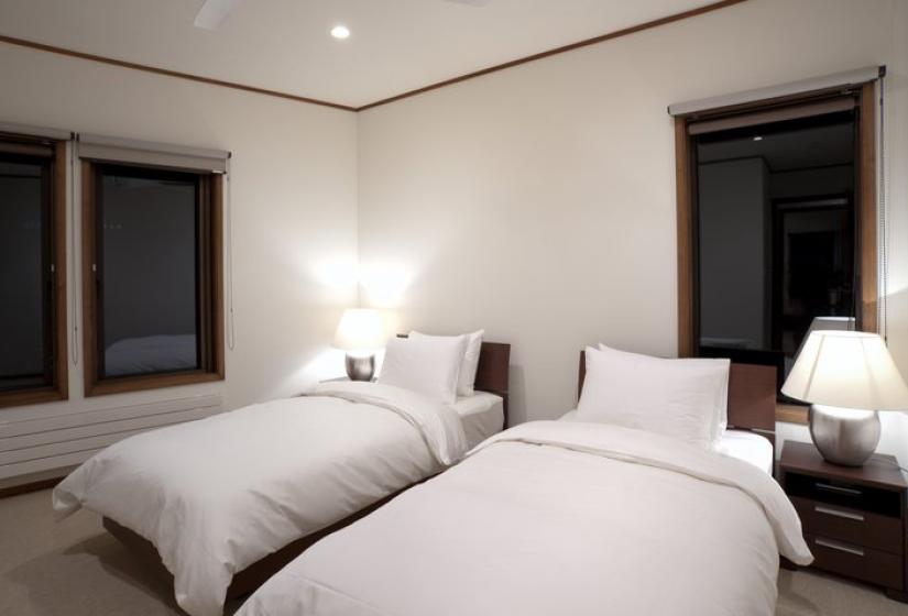 Modern twin bedroom with two white beds, bedside table with lamp, minimalist design, and dark-framed windows in Chalet Snowbird.