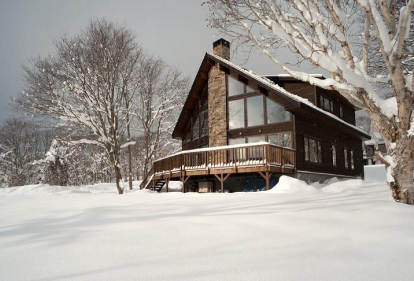 Wooden chalet with large windows surrounded by deep snow and bare winter trees against a misty mountain backdrop in Niseko.