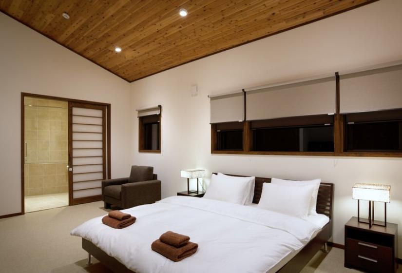 Modern bedroom with wooden ceiling, king-sized bed with white linens, bedside tables with lamps, minimalist Japanese-inspired design, and sliding doors.