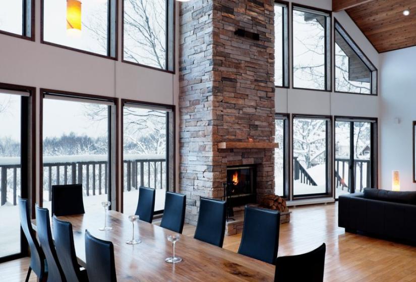Spacious dining area with high vaulted wooden ceiling, floor-to-ceiling windows, stone fireplace, wooden dining table with black chairs, and views of snowy mountains outside.