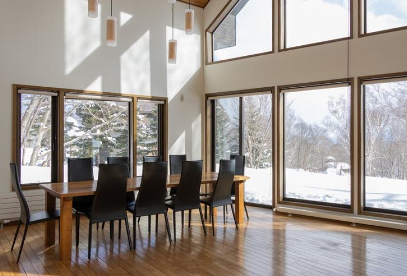 Spacious dining area with wooden floor, large windows offering snow-covered mountain views, high ceiling, and elegant dark dining chairs around a long wooden table.