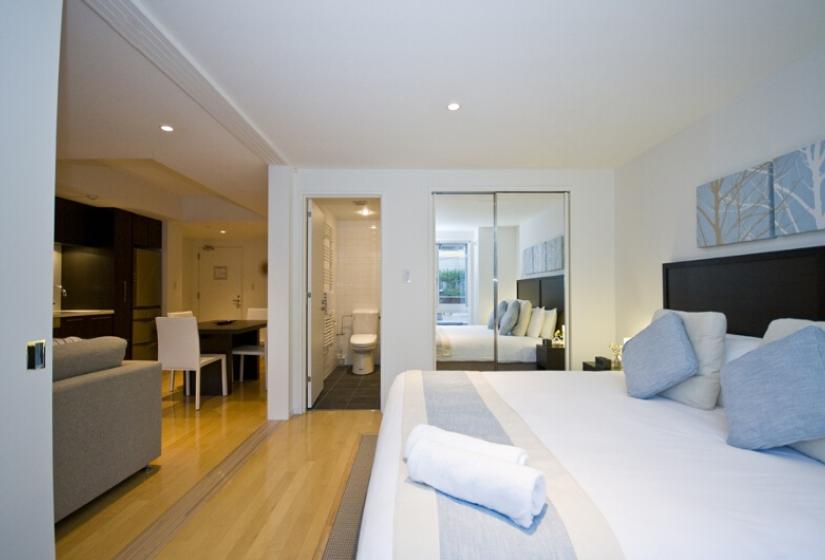 Alpen Ridge Niseko 1 bedroom apartment featuring stylish master bedroom with king bed blue accent pillows light wooden flooring connecting bathroom door kitchenette view and bright contemporary design for couples ski accommodation